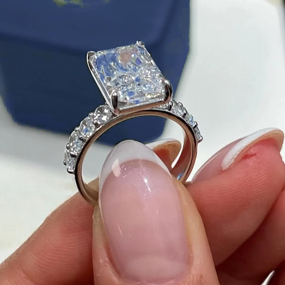 Fingertips holding a radiant-cut lab-grown diamond engagement ring with a diamond band, a navy ring box behind