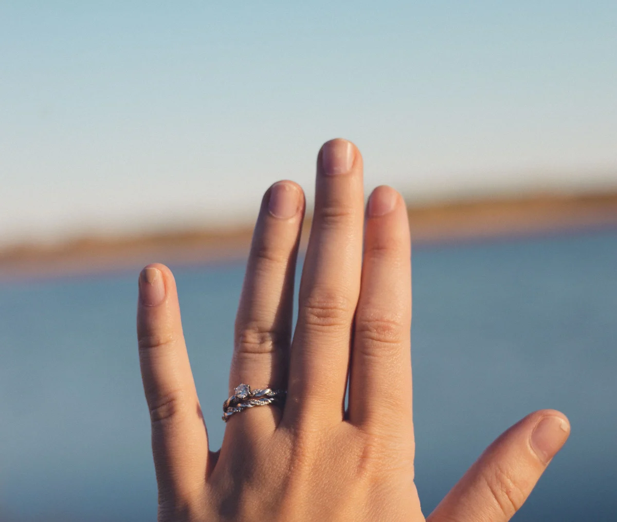 A woman’s hand raised against a blue lake at golden hour, wearing a twisted lab-grown diamond ring
